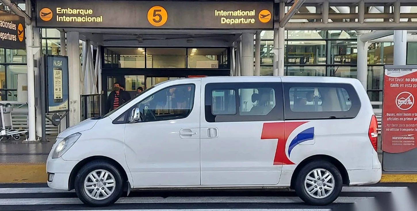 Van blanca de Transfer Chile, con el logo rojo y azul, frente al acceso de Embarque Internacional 5 del aeropuerto de Santiago